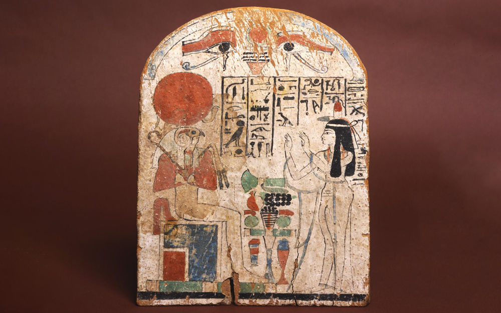 Round topped wooden stela belonging to the singer of Amun, Tasheryt. She stands on the right side of the stela wearing a long, sheer dress and has her hands raised in adoration to Re-Horakhty who is seated on the left. 